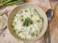 Kremowe risotto ze szparagami, posypane parmezanem, z trzema zielonymi szparagami na wierzchu. Obok miseczka z tartym serem i pęczek świeżych szparagów.
