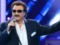 Krzysztof Krawczyk w okularach przeciwsłonecznych i granatowej marynarce śpiewa na scenie, gestykulując prawą ręką.