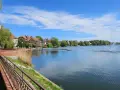 Spokojne jezioro zamkowe z fontanną i drewnianym pomostem, otoczone zielenią i zabudowaniami.