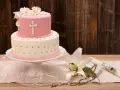 Elegancki dwupoziomowy tort na chrzest w różowo-białych barwach, ozdobiony krzyżem i kwiatami. Obok lilia, różaniec i świeca.