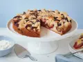Ein einfacher Pflaumenkuchen mit Streuseln auf einem Tortenständer, daneben eine Schüssel Schlagsahne und ein Tortenheber.