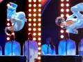 Wokalista w błyszczącym niebieskim stroju wykonuje akrobacje na scenie podczas gali nagród Grammy. Laureaci nagród grammy w białych garniturach stoją za fortepianami.
