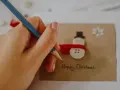 Ręka z ołówkiem pisze "Happy Christmas" na kartce z bałwankiem z guzików. Idealny przykład, jak narysować kartkę świąteczną.