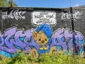 Enkel graffiti på en tegelvägg visar en brun nallebjörn i blå overall och mössa, med texten "Utan blommor ingen honung!".