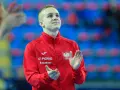 Adam Jędraszek w czerwonej kurtce z orłem, z dłońmi uniesionymi w geście radości lub powitania.