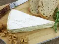 Tranche de fromage à croute lavée crémeux, accompagnée de pain, noix et herbes aromatiques sur une planche en bois.