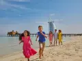 Rodzina cieszy się słonecznym dniem na plaży w Dubaju. Dzieci biegną w stronę morza, a w tle widać słynny hotel Burj Al Arab.