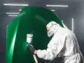Person i skyddsdräkt lackar en grön motorhuv med sprutpistol i en verkstad.