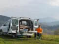Mężczyzna popija kawę przy rozłożonym stoliku obok swojego białego Dacia campera, rozkoszując się widokiem gór.
