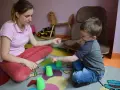 Terapia behawioralna autyzm: chłopiec i terapeutka bawią się kubeczkami na kolorowym dywanie.