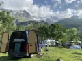 En campingplats med flera husbilar, inklusive en blå med öppna bakdörrar, i en dal med snöklädda berg i bakgrunden. En perfekt plats för äventyr med edins husbilar.