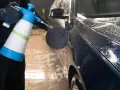 En person med svarta handskar sprutar skum på en mörk metallicbil med en blå och vit sprayflaska. Bilvax metallic ger en glansig finish.