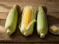 Drei Maiskolben liegen auf Holz. Einer ist geschält und zeigt die gelben Körner. Daneben eine kleine Schale mit Butter und Salz – bereit, um Mais zu kochen.