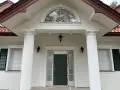Elegancka fasada domu z białymi pilastrami, ozdobnym oknem nad drzwiami i stylowymi lampami.