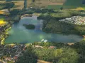 Luftaufnahme des Feldmarksees Sassenberg mit einer kleinen Insel, Booten am Steg, einem Wohngebiet und Feldern.