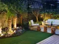 Un jardín poco mantenimiento con césped artificial, palmeras iluminadas y una zona lounge con sofá blanco y sillón.