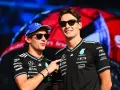 Dos pilotos de Fórmula 1, George Russell y un compañero, sonríen con gafas de sol. Russell lleva una gorra azul y ambos visten camisetas negras con logotipos de patrocinadores.
