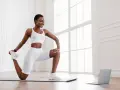 Femme en tenue de sport blanche effectuant un exercice quadriceps sur un tapis, guidée par un ordinateur portable.