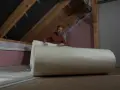 Mężczyzna w rękawiczkach przygotowuje ocieplenie dachu, rozwijając dużą rolkę izolacji.