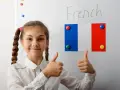 Uśmiechnięta dziewczynka z warkoczami pokazuje kciuki w górę na tle tablicy z napisem "French" i flagą Francji. To symbol pozytywnego podejścia do nauki, być może w ramach systemu edukacji we Francji.