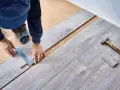 Ręce majsterkowicza mierzą i zaznaczają panele winylowe do cięcia. Młotek i kawałek podłogi leżą obok.