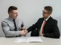Dwóch mężczyzn w garniturach ściska sobie dłonie nad stołem, na którym leży dokument. To symboliczne podpisanie umowy przedwstępnej.