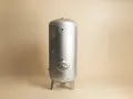 En silverfärgad hydrofor eller hydropress står på tre ben mot en ljusbeige bakgrund.