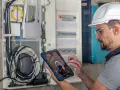 Technik w kasku i okularach ochronnych sprawdza przegląd instalacji elektrycznej domu na tablecie. Widoczne przewody i rozdzielnica.