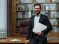Uśmiechnięty dyrektor szkoły w garniturze, trzymający laptopa, stoi przy biurku w gabinecie.