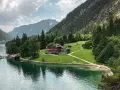 Gaisalm am Achensee: ein Gasthaus mit roten Fensterläden, umgeben von grünen Wiesen und Bergen. Ein Schiff liegt am Steg.