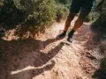 Jambes d'un homme en short et chaussures de trail, s'engageant sur un sentier rocailleux et poussiéreux. L'ombre de son corps s'étire sur le chemin, suggérant un rythme de fast hiking sous le soleil.