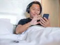 Mężczyzna w słuchawkach leży w łóżku, korzystając z telefonu. To prosty sposób, jak słuchać audiobooka na telefonie, relaksując się.