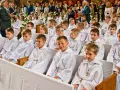 Dzieci w bieli, gotowe na rocznicę komunii świętej, siedzą w kościele. W tle widać zgromadzonych gości i fotografa.
