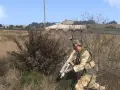 Żołnierz w kamuflażu z karabinem ukryty za krzakami, w tle pojazd opancerzony.
