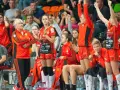 Drużyna piłki ręcznej Metraco Zagłębie Lubin w pomarańczowych strojach, z trenerką i zawodniczkami na ławce rezerwowych.