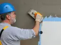 Budowlaniec w niebieskim kasku nakłada tynk na styropian. Prace nad fasadą, w tym suche tynki, postępują.