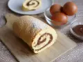 Un delizioso rotolo alla nutella, spolverato di zucchero a velo, pronto per essere gustato. Ingredienti freschi, uova e crema al cioccolato, completano la scena.