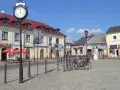 Słoneczny stary rynek łowicz z brukowanym placem, budynkami z czerwonymi dachami i zegarem na słupie.