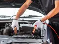 Hands in white gloves connect jumper cables to a car battery. This process helps answer how long does it take to charge a car battery.