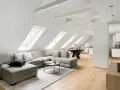 Soggiorno mansardato con divano angolare grigio, tavolino nero e travi a vista moderne dipinte di bianco che creano un'atmosfera luminosa.