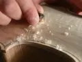 Hände bearbeiten mit einem Werkzeug das helle Holz einer Geige, Späne fliegen. Die Frage "aus welchem Holz besteht die Geige" wird hier visuell beantwortet.