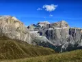 Vaste cime rocciose del gruppo Sella Dolomiti sotto un cielo azzurro con nuvole bianche. Prati verdi e sentieri serpeggiano sui pendii.