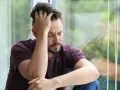 Mann sitzt mit geschlossenen Augen und der Hand am Kopf vor einem Fenster.