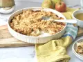 Ein goldbrauner Apfel-Crumble in einer Auflaufform, bereit zum Servieren. Daneben eine Tasse Tee und frische Äpfel.