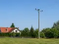 Słup na działce, obok domu z czerwoną dachówką i żywopłotem. W tle zielone drzewa i błękitne niebo.
