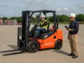 Pomarańczowy wózek widłowy Noblelift z operatorem w kamizelce i kaskiem, obok stoi mężczyzna z notatnikiem.