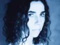 PJ Harvey, z kręconymi włosami, patrzy intensywnie. Jej twarz, w odcieniach niebieskiego, zdaje się opowiadać historię z "I Inside the Old I Dying".