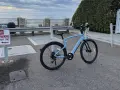 A light blue electric city bike is parked on asphalt near a "No Parking" sign, with the ocean and cloudy sky in the background.