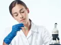 Giovane tecnologo alimentare in camice bianco, con guanti blu, analizza un campione con un contagocce accanto a un microscopio.