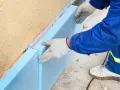 Ręce w rękawiczkach układają niebieski styropian przy ocieplanej ścianie fundamentowej.
